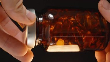 Karanlık bir zemin karşısında Omega 3 kapsülleriyle dolu kahverengi cam bir şişeyi döndürerek yüzeydeki ışık yansımalarını yakalıyoruz.