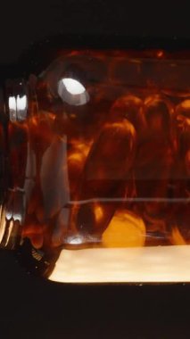 Dikey video. Karanlık bir zemin karşısında Omega 3 kapsülleriyle dolu kahverengi cam bir şişeyi döndürerek yüzeydeki ışık yansımalarını yakalıyoruz.