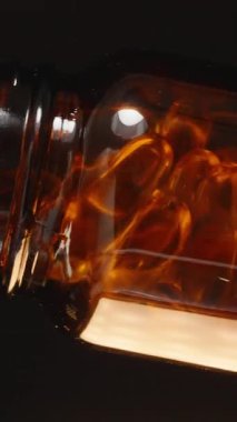 Dikey video. Omega 3 kapsüllerini kahverengi bir cam şişeden açık bir avuç içine dökerek karanlık bir arkaplan yakalama hareketi