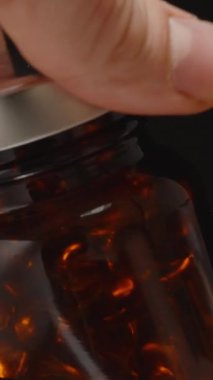 Dikey video. Metal Lid 'i kaldırarak Kahverengi Cam Omega 3 Kapsülünü Açan Eller, İçerideki Kapsülleri Karanlık Bir Arkaplana Karşı Gösteriyor
