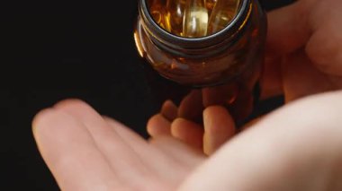 Omega 3 kapsüllerini, kahverengi cam bir şişeden açık bir avuç içine dökerek, karanlık bir arkaplana karşı hareketi yakalıyor.
