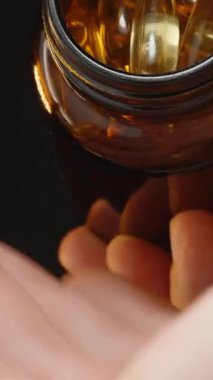 Dikey video. Omega 3 kapsüllerini, kahverengi cam bir şişeden açık bir avuç içine dökerek, karanlık bir arkaplana karşı hareketi yakalıyor.
