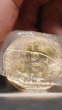 Dikey video. Elle tutulan Bitcoin Isıtılmış Metal bir yüzeyde kısmen erimiş bir buz küpü içinde, yükselen buharla, finansal belirsizliği ve piyasa istikrarsızlığını temsil eden