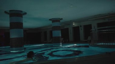 Gece Büyük Kapalı Havuzda Yüzen Kadın, Karanlık Başlıklı Sütunlar ve Salon Sandalyeleriyle çevrili, Geniş Açı Bakış Açısı, Huzur Sağlığı Konsepti