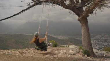 Arkaplanda Bulutlu Gök ve Dağ Kıyısındaki Bir Ağaçta Sallanan Kadın, Yan Açı, Özgürlük ve Macera Konseptinden Yakalandı.