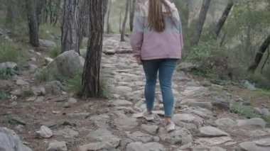 Pembe ve gri ceketli, kot pantolonlu ve kepli bir kadın uzun ağaçlar ve yeşilliklerle çevrili kayalık bir orman yolunda yürüyor doğayı keşfediyor ve yokuş yukarı tırmanıyor.