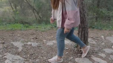 Yoğun bir ormanda engebeli bir toprak patikada yürüyüş yapan bir kadın uzun ağaçlar ve yemyeşil bitkilerle çevrili bir kep, pembe ceket ve kot pantolon giyerken kayaların üzerinden atlıyor.