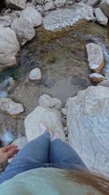 Dikey video. Dalgalı Nehirde Büyük Kayaların Üzerinde Dolaşan Kadın, İstikrar Arazisi 'ni dikkatle tutarken Kayalıklar ve Vahşi Doğa, İlk Perspektif, 360 derecelik Kamerayla çevrili