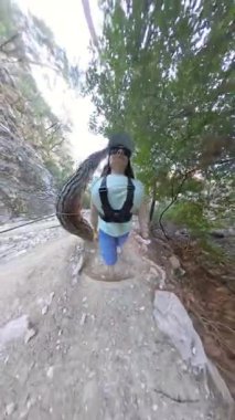 Dikey video. Kanyonda Rocky Yolu 'nda diz çökmüş, güneş gözlüğü ve kamera kayışları takan, Ağaçlarla çevrili ve çarpık bir ağaç gövdesi olan bir kadının küçük gezegen efekti, 360 derecelik kamera.