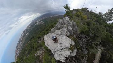 Rocky Cliff 'in kenarında oturan bir çift 360 derecelik Küresel Panorama' da Uzak Deniz ve Şehre hayranlık duyuyor.
