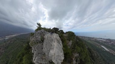 Denize Bakan Büyük Rocky Ledge 'de oturan bir çift ve Geniş Açı Görüş Alanında Bulutlu Gökyüzü Altında Uzak Bir Şehir Olan Yeşil Vadi
