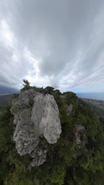 Dikey video. Denize Bakan Büyük Rocky Ledge 'de oturan bir çift ve Geniş Açı Görüş Alanında Bulutlu Gökyüzü Altında Uzak Bir Şehir Olan Yeşil Vadi