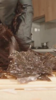 Dikey video. Kamera, Siyah Eldiven Giyen El Gibi Yakın Çekimde İlerliyor. Gözyaşları Kurumuş Nori Yaprakları Ahşap Yüzeyde Bulanık Mutfak Arkaplanlı