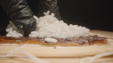 Kamera, Siyah Eldiven Giyen El Olarak Nori Çarşafı boyunca ilerler. Suşi Pirincini nazikçe yayar, Doku ve Detaylara Odaklı Makro Yakın plan Çeker.
