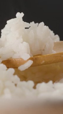 Dikey video. Tahta kaşık kaldırma Buharlı Taze Pişmiş Beyaz Pirinç Kaseden, Yumuşak Doku ve Nemlendirmeyi Gösteren Karanlık Bir Arkaplana Karşı Sıcak Buharda Yükseliş