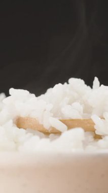 Dikey video. Bir kase taze pişmiş beyaz pirinç üzerine yükselen buhar bir tahta kaşık gibi yumuşak ve nemli tahıllar sıçrıyor, sıcak ve rahatlatıcı bir sahne yaratıyor