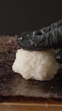 Dikey video. Siyah Eldivenli Eldivenler Nori çarşafı üzerinde Yapışkan Beyaz Pirinç 'i, Bambu Mat' ın Üzerinde, Yuvarlanan Suşi İçin Üs 'ü Hazırlıyor