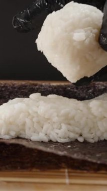 Dikey video. Siyah Eldivenli Yapışkan Beyaz Pirinçli Kalın bir katmanı Nori çarşafına bastırıp Suşi Roll 'u özenle ve titizlikle hazırlıyor.