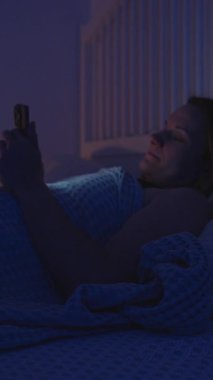 Dikey video. Kadın Yatakta Tarafına Dönüyor ve Uykuya Dalarken Akıllı Telefonu Yatıyor Ekran, Yan Görünüm, Yatma Zamanı Düzenleme Konsepti