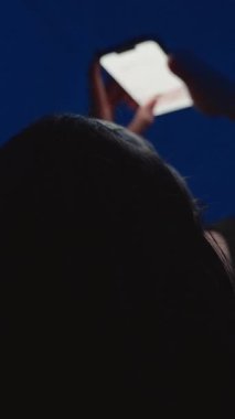 Dikey video. Loş Mavi Işık Odası 'nda Sırtüstü Uzanmış Bir Elle Arkadan Başının Arkasına Uzanıp Akıllı Telefonu Tutan Kadın, Gece Bakışları Konsepti