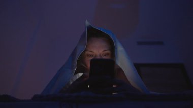 Sim Room 'da Battaniyenin Altındaki Kadın Düşük Açı' dan Yüzünde Ekran Işığı olan Akıllı Telefona Bakıyor, Gizli Bakış Konsepti