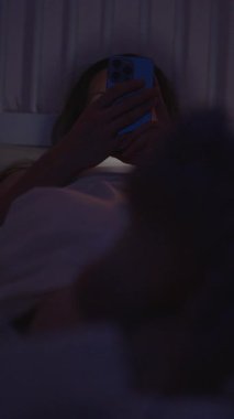Dikey video. Kadın Smartphone 'a Bakar Ayaklarıyla Yatakta Uzanırken Kameraya doğru, Loş Aydınlatma Odasında, Yakın-Yakın-Düşük Açı, Rahatlatıcı Akşam Konsepti
