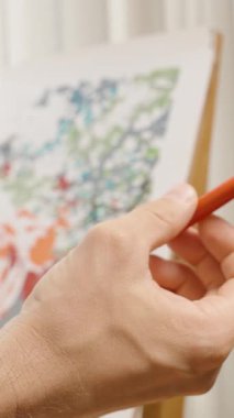 Dikey video. Arkaplanda Soyut Tuval ile Parmaklar Arasında Zarif Bir Uzun İnce Boya Fırçası Döndüren Sanatçı ve Bir Pencereden Yumuşak Işık, Alanın Sığ Derinliği, Sanatsal Kontrol Konsepti