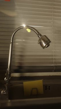 Dikey video. Güneş Işıkları Kapalı Pencere Aralıklarından Gizlice Bakıyor Mutfak Lavabosunun Üzerinde Esnek Musluk ve Cam Yakınında, Göz Seviyesi Açısı, Geç Öğlen Işığı Konsepti