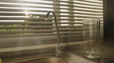 İçme Suyu Yavaşça Süzgeç Musluğundan Lavabo 'ya Damlıyor. Yanında duran Cam ve Sunlight Perdelerden Geçiyor, Göz Seviyesi Açısı, Su Koruma Konsepti