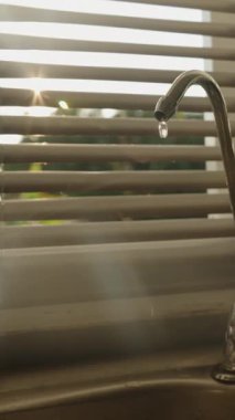 Dikey video. Süzgeç musluğundan damlayan su El, Valfı Sıkıca Kapatmak İçin Döndürürken, Güneş Işığı Perdelerden, Göz Seviyesi Açısı, Su Tasarruf Konsepti