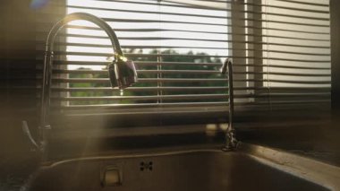 Su yavaşça Esnek Musluktan Günbatımında Batımına akıyor. Pencere Perdelerinden Altın Işık, Göz Seviyesi Açısı, Su Atık Kavramı
