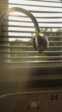 Dikey video. Güneş Işığı Perdelerden Süzülürken Esnek Musluktan Damlayan Su Atık, Göz Seviyesi Açısı, Su Koruması Konseptini Engellemek İçin Sıkıca Kapanıyor