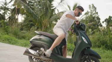 Beyaz Gömlekli Adam Yeşil Scooter Bağlıyor ve Arka planda Palmiye Ağaçları, yandan görüş, Seyahat ve Özgürlük Konsepti ile Tropikal Yol Uzağında Sürüyor
