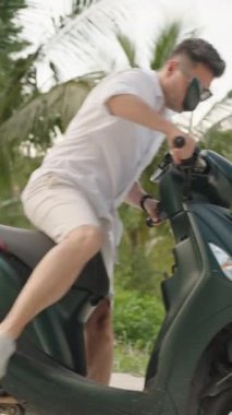 Dikey video. Beyaz Gömlekli Adam Yeşil Scooter Bağlıyor ve Arka planda Palmiye Ağaçları, yandan görüş, Seyahat ve Özgürlük Konsepti ile Tropikal Yol Uzağında Sürüyor