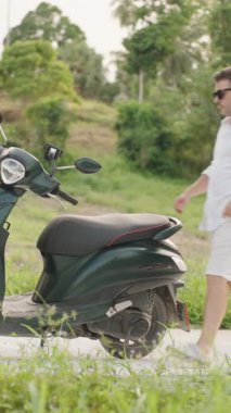 Dikey video. Beyaz Giysili Adam Koyu Yeşil Scooter Kırsal Yol 'da, Ağaçlar ve Çimenli, geniş açılı, Tropikal Keşif Konsepti