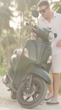 Dikey video. Beyaz tişörtlü ve şortlu adam Scooter 'a yaklaşıyor ve Virajlı Tropikal Yol' da Depo Bölümü açıyor, düşük açılı görüş, Pratik Seyahat Anı Kavramı