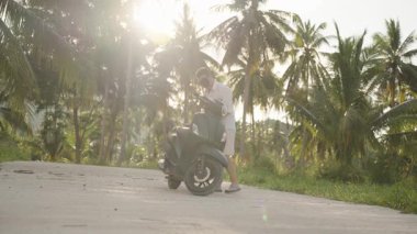Man Walking to Scooter and Accessing Bagk Before Leave on Curved Tropical Road with Sunset Light, geniş açılı güneş parlaması efekti, Spontane Seyahat Anı kavramı