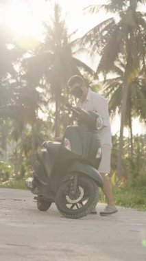 Dikey video. Man Walking to Scooter and Accessing Bagk Before Leave on Curved Tropical Road with Sunset Light, geniş açılı güneş parlaması efekti, Spontane Seyahat Anı kavramı