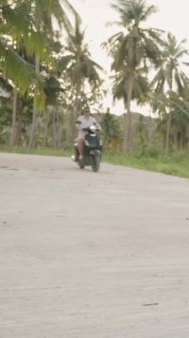 Dikey video. Beyaz gömlekli adam, Scooter 'la Tropikal Ada' daki Boş Beton Yolu 'nda Palm Trees ve Hills' te, geniş açılı manzaralı, Ada Özgürlük Konsepti.