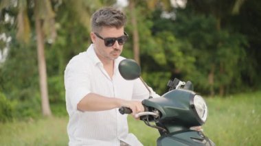 Kamera Sürücüden Arka plandaki Palm Trees 'e geçerken, Beyaz Gömlekli Adam Scooter Başlıyor. Derinlik kayması ve Tropikal Ayrılış Konsepti ile yakın plan.