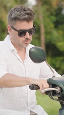 Dikey video. Kamera Sürücüden Arka plandaki Palm Trees 'e geçerken, Beyaz Gömlekli Adam Scooter Başlıyor. Derinlik kayması ve Tropikal Ayrılış Konsepti ile yakın plan.