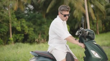 Beyaz tişörtlü adam scooter 'da oturuyor ve Lush Tropical Setting' de ileriye bakıyor, orta yan görüş, Beklenti ve Seyahat Konsepti