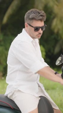 Dikey video. Beyaz tişörtlü adam scooter 'da oturuyor ve Lush Tropical Setting' de ileriye bakıyor, orta yan görüş, Beklenti ve Seyahat Konsepti