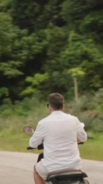 Dikey video. Arka Manzaradaki virajlı bir yol boyunca yoğun yeşil alandan geçen Scooter 'lı adam, Kalın Bitkiler ve Kamu Hizmetleri Kutupları ile çevrili, Ada Seyahat Konsepti