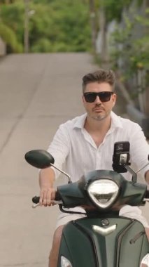 Dikey video. Beyaz Gömlekli Adam ve Güneş Gözlüklü Adam Yeşil Scooter ile Banliyö Yolu 'nda Çit ve Ağaçlarla çevrili Kameraya doğru, Merkezli Kafadan Görüntü, Şehir Hareketi Konsepti