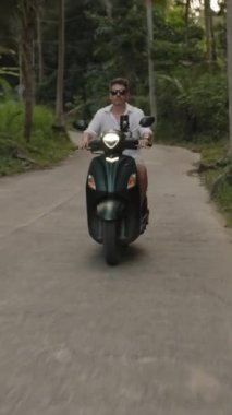Dikey video. Scooter 'ı Orman Yolu' ndaki kameraya doğru süren adam yemyeşil sebzeli, far açık frontal geniş açılı, Eco Travel Concept