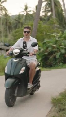Dikey video. Uzun Palmiye Ağaçları ve Uzak Tepeler, İleri Hareketli Hafif Açı, Tropikal Macera Konsepti ile çevrili Scooter Çevresindeki Adam