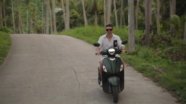 Beyaz gömlekli adam ve Dar Palmiye Ağaçları Arasında Scooter süren güneş gözlüklü adam, Yavaş Çekimde Frontal Manzara, Tropikal Yolculuk Konsepti