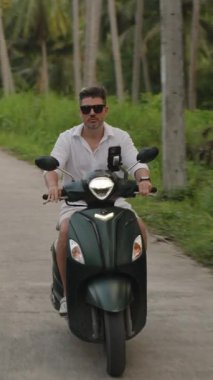 Dikey video. Beyaz gömlekli adam ve Dar Palmiye Ağaçları Arasında Scooter süren güneş gözlüklü adam, Yavaş Çekimde Frontal Manzara, Tropikal Yolculuk Konsepti