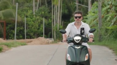 Beyaz Gömlekli Adam Viraj Yolu 'nda yavaşça scooter kullanıyor. Palmiye ağaçlarıyla aynı hizada. Önden hafif açılı, Yavaş Hareketli, Tropikal Sürücü Konsepti.